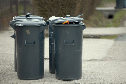 Two large, dark grey wheeled wheelie bins with closed lids are positioned on a paved outdoor driveway, likely in a residential or urban area. The bin on the left has a smooth surface with a small rectangular indentation on the front, while the bin on the right has its lid slightly ajar, revealing an orange item inside. Both bins appear to be made of durable plastic and are designed for rubbish collection, with the left bin’s lid fully secured and the right bin's lid partially open. The background shows a blurred view of a pathway or road with a slight incline, some greenery, and a small bridge or culvert in the distance, suggesting a suburban environment. The scene is lit with natural light, indicating daytime, and the overall setting aligns with waste disposal services in the Shadwell area. The scene exemplifies typical rubbish collection preparations managed by services like Office Clearance Shadwell.