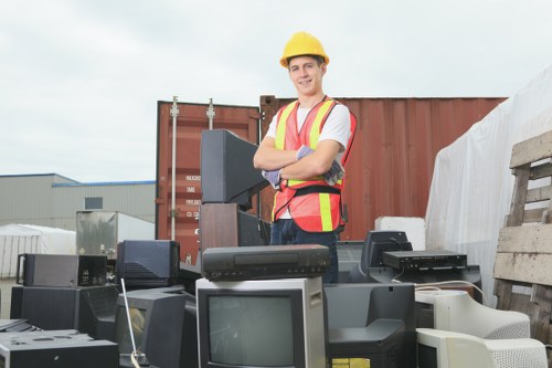 A young male worker wearing a yellow safety helmet and a high-visibility vest, standing outdoors amidst a variety of discarded electronic items including old computer monitors, CRT screens, and computer towers, with a large shipping container and industrial warehouses visible in the background. The electronic waste is haphazardly arranged on a surface, indicating a collection or clearance process. The scene is captured under natural daylight, with a neutral sky overhead. This image illustrates a rubbish removal or clearance operation in an industrial or commercial setting, showcasing the type of waste managed by Office Clearance Shadwell, which is involved in removing bulky and electronic rubbish from premises within the Shadwell area, close to London's East End. The variety of outdated electronics highlights the types of waste handled during such clearance services, emphasizing professional disposal of electronic and general rubbish.