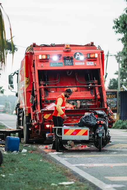 A large, red, metal rubbish skip with visible rust and chipped paint is positioned against the corner of a modern building, with its side leaning slightly into a narrow outdoor alley. Around the base