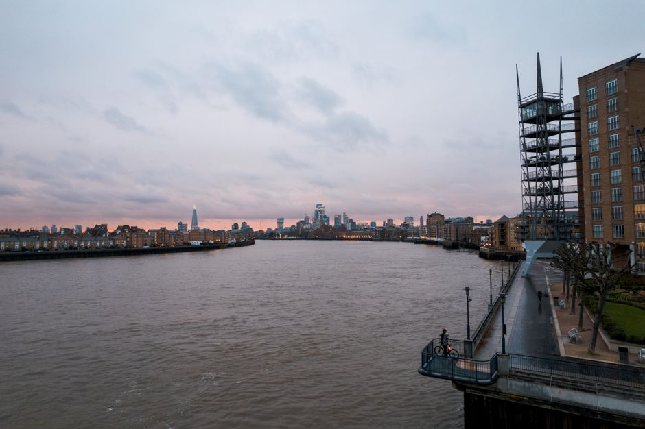 A view of an urban river scene during daylight shows a calm waterway with gentle ripples in the foreground, likely caused by a passing boat. Along the riverbank on the left side, there are several mod