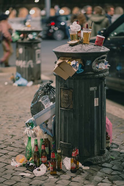 A black metal public trash bin situated on a cobblestone street corner, with an overflowing assortment of rubbish surrounding its base. The lid of the bin is slightly ajar, and the top surface is clut